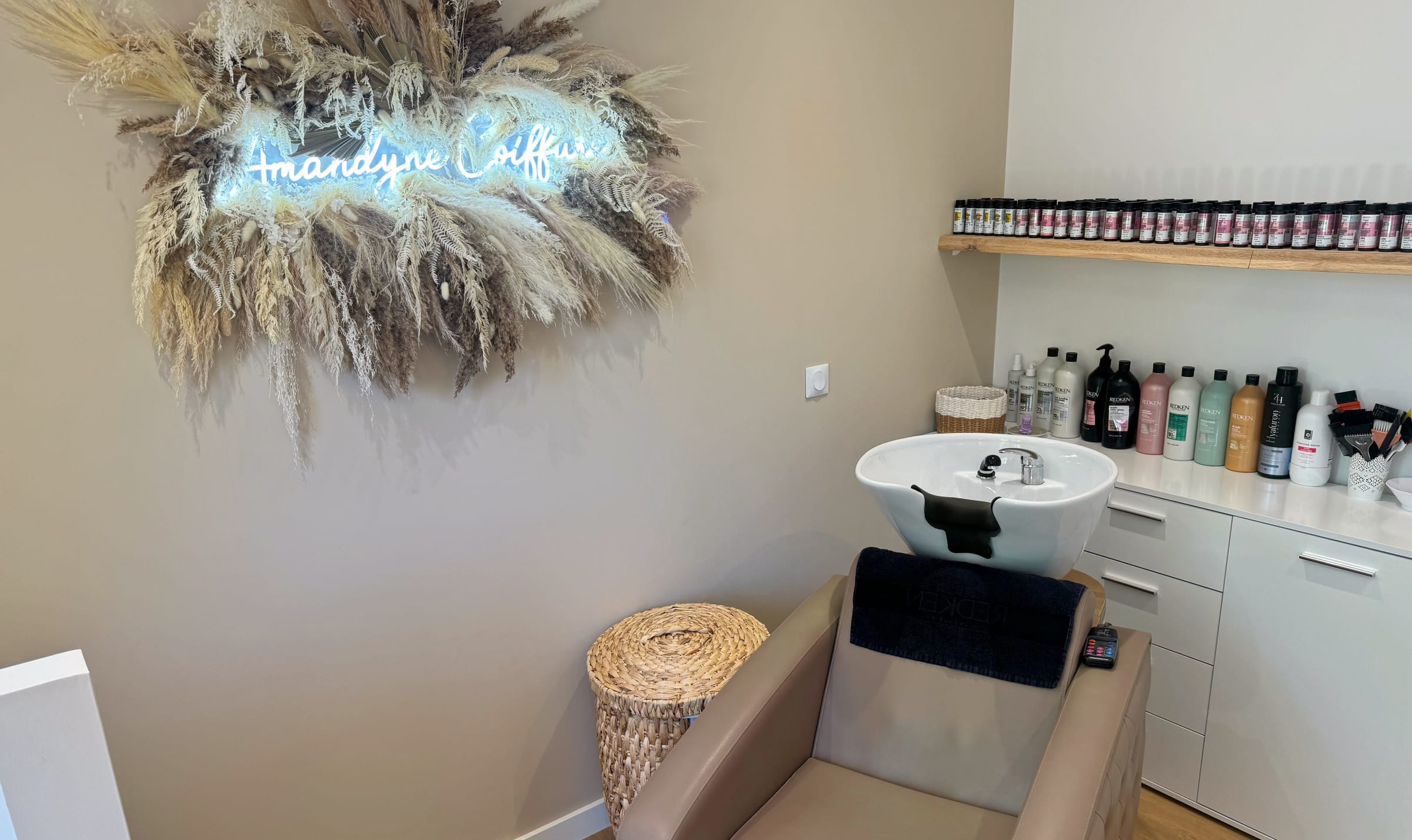 Le néon « Amandyne Coiffure » entouré d’une couronne de fleurs séchées, au-dessus du bac à shampoing